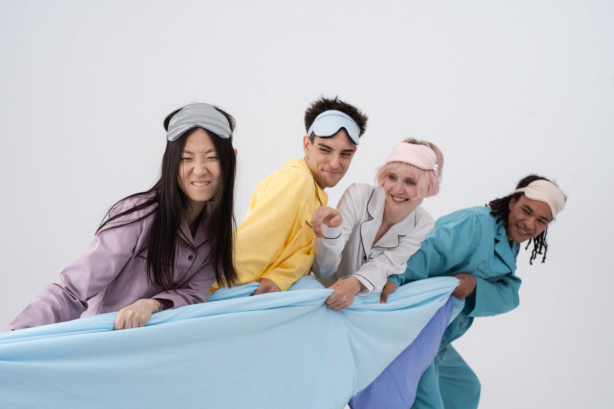 Four friends in pajamas and sleep masks pulling a light blue sheet